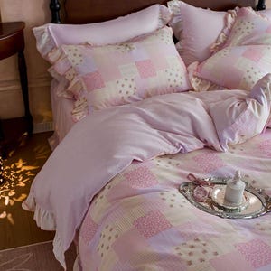 Puede incluir: Una cama con un edredón de patchwork rosa y blanco y fundas de almohada a juego. El edredón presenta una mezcla de patrones florales y geométricos. Las fundas de almohada tienen bordes con volantes. Una bandeja plateada con una vela y objetos decorativos está sobre el edredón.