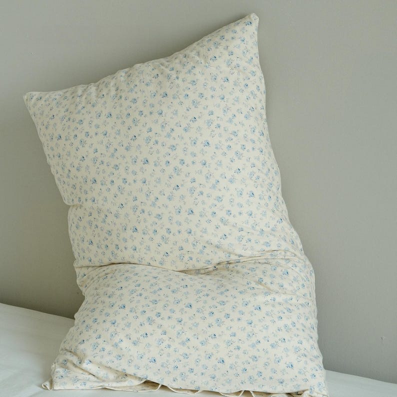 May include: A large, rectangular pillow with a cream-colored background and a pattern of small blue floral designs. The pillow is slightly crumpled, suggesting a soft, comfortable texture, and is placed on a white surface.