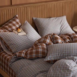 Pode incluir: Uma cama com cabeceira de madeira e roupa de cama com um padrão xadrez marrom, azul e branco. O conjunto inclui travesseiros, um edredom e um lençol com elástico. Uma pequena vela está em cima de um travesseiro.