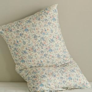 May include: A large, square pillow with a floral pattern. The pillow features a cream-colored background with a repeating pattern of small blue and red flowers. The pillow is soft and appears to be made of fabric.