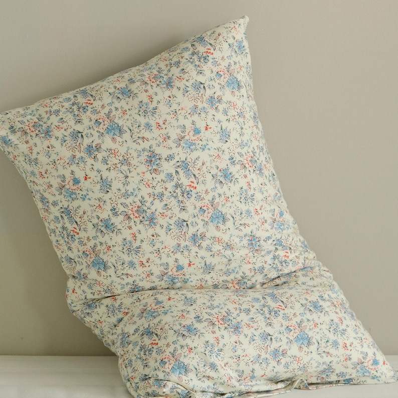 May include: A large, square pillow with a floral pattern. The pillow features a cream-colored background with a repeating pattern of small blue and red flowers. The pillow is soft and appears to be made of fabric.