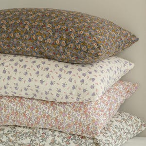 May include: A stack of four rectangular pillows with floral patterns. The top pillow has a gray background with yellow and white flowers. The second pillow has a white background with purple flowers. The third pillow has a pink background with orange flowers. The bottom pillow has a white background with pink flowers.