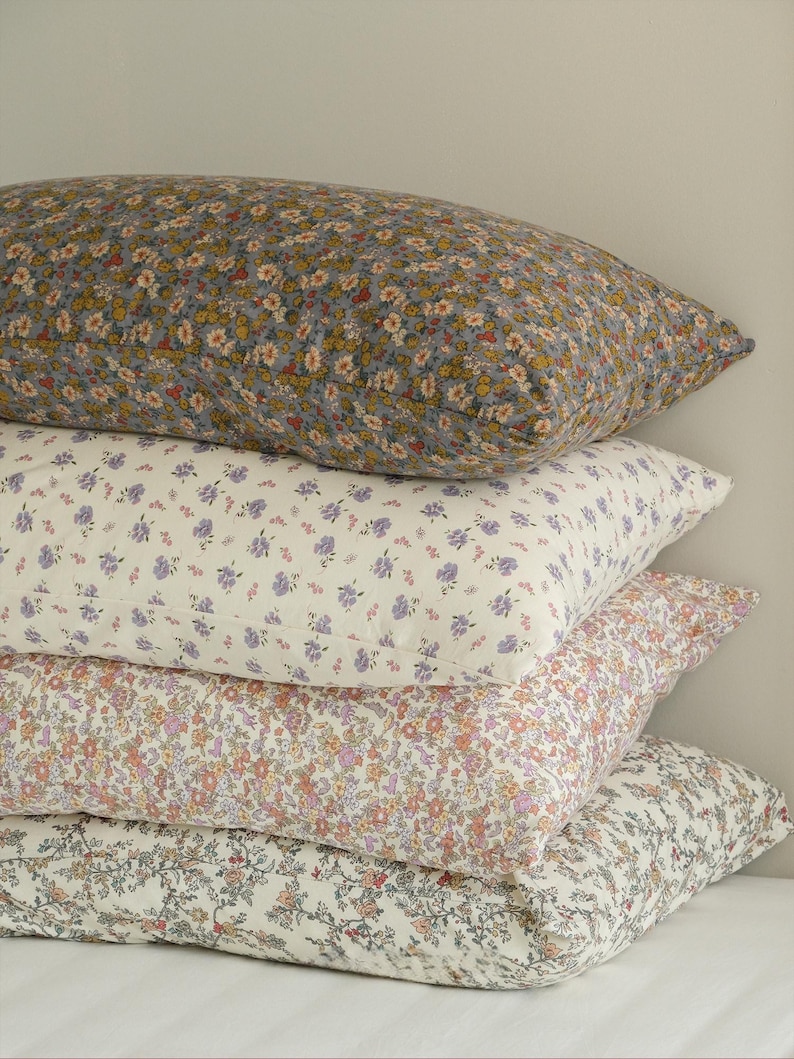 May include: A stack of four rectangular pillows with floral patterns. The top pillow has a gray background with yellow and white flowers. The second pillow has a white background with purple flowers. The third pillow has a pink background with orange flowers. The bottom pillow has a white background with pink flowers.