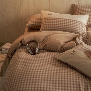 Peut inclure: Un lit avec une housse de couette et des taies d'oreiller à carreaux marron et blancs. Le lit a une couette marron et plusieurs oreillers. Un casque marron repose sur la couette. Les oreillers ont un petit motif d'ours.