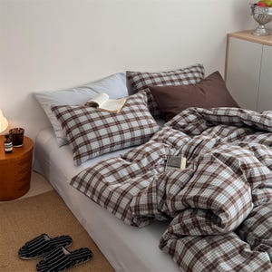 May include: A bed with a brown and white plaid comforter, pillowcases, and pillows. A book is open on one pillow. A small wooden side table with a lamp and a bottle is to the left of the bed. Striped slippers are on the floor.