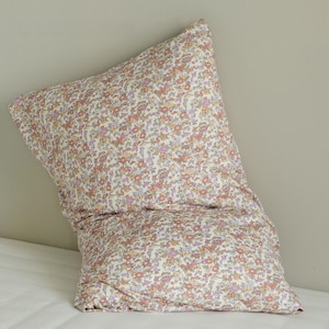 May include: A floral patterned pillowcase in a cream color with small orange, pink, and purple flowers. The pillowcase is folded and resting on a white surface, with a neutral-colored wall in the background.