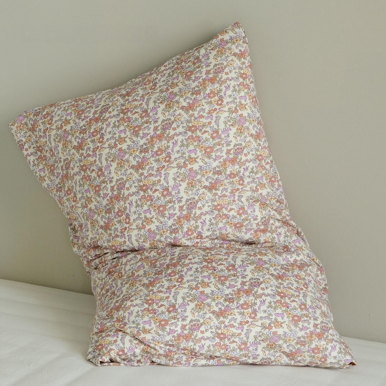 May include: A floral patterned pillowcase in a cream color with small orange, pink, and purple flowers. The pillowcase is folded and resting on a white surface, with a neutral-colored wall in the background.