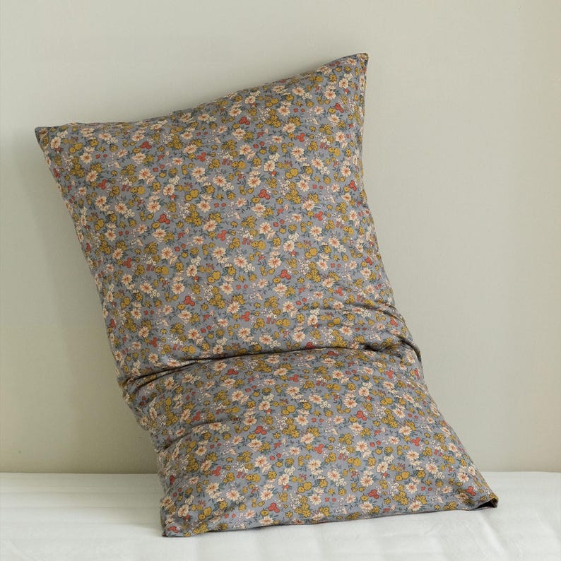 May include: A large, rectangular pillow with a floral pattern. The pillowcase is a muted gray color, covered in a repeating pattern of small yellow, orange, and white flowers. The pillow is resting on a white surface.