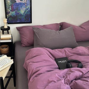 May include: A bedroom scene featuring a bed with mauve bedding, including pillows and a duvet. A book with "PRADA CATWALK" on the cover and black headphones rest on the bed. A framed art print hangs on the wall, and a small side table is visible.