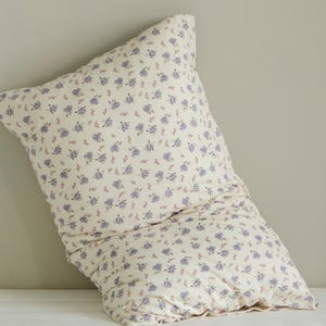 May include: A cream-colored pillow with a repeating pattern of small purple and pink flowers. The pillow is soft and appears to be made of cotton or a similar fabric. The floral design adds a touch of elegance to the bedding.
