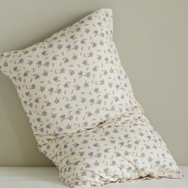 May include: A cream-colored pillow with a repeating pattern of small purple and pink flowers. The pillow is soft and appears to be made of cotton or a similar fabric. The floral design adds a touch of elegance to the bedding.