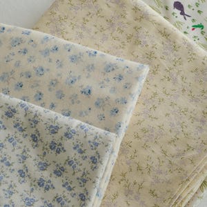 May include: A collection of folded fabric pieces featuring floral and botanical patterns. The fabrics display a variety of designs, including small blue flowers on a cream background, and sprigs of lavender and green on a light yellow background.