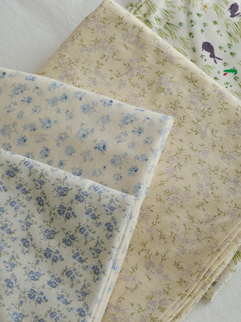 May include: A collection of folded fabric pieces featuring floral and botanical patterns. The fabrics display a variety of designs, including small blue flowers on a cream background, and sprigs of lavender and green on a light yellow background.