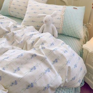 May include: A bed with floral bedding and a stuffed bunny. The white bedding has blue floral patterns and is paired with pillows featuring a gingham pattern and lace trim. The overall color scheme is soft and inviting, with a focus on comfort and a touch of vintage charm.