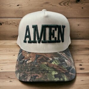 May include: A beige baseball cap with the word "AMEN" embroidered in dark green block letters. The brim features a camouflage pattern in shades of brown, green, and tan. The cap is set against a wooden background.