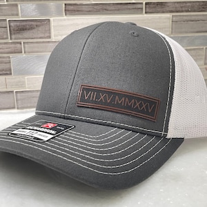 May include: A gray trucker hat with a white mesh back. The front panel features a brown leather patch with Roman numerals. The hat has a curved brim and white stitching details. The brand tag is visible on the side.