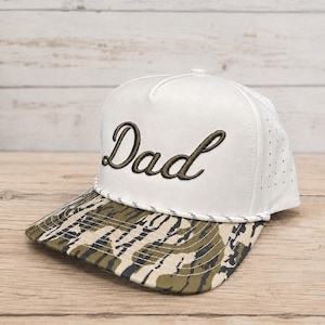 May include: A white baseball cap featuring the word Dad embroidered in dark green script on the front panel. The cap includes a white braided cord across the base of the crown and a brim with a green, brown, and tan camouflage pattern.