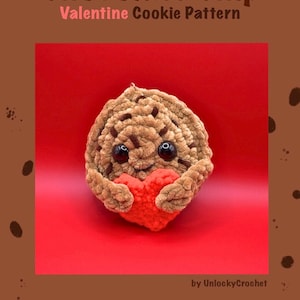 Könnte beinhalten: Ein gehäkelter Schokoladen-Chip-Keks, der ein rotes Herz hält. Der Keks hat schwarze Knopfaugen und ist aus braunem Garn mit dunkleren braunen Schokoladenstückchen gefertigt. Der Text "Chocolate Chip Valentine Cookie Pattern" steht oben.
