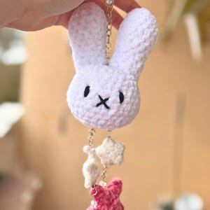 Könnte beinhalten: Ein weißer Plüsch-Hasen-Schlüsselanhänger mit schwarzen Augen und einem gestickten 'X'-Mund. Darunter hängen Charms in Form von weißen und rosa Sternen und einer Tropfenform, alle verbunden durch eine silberne Kette. Der Schlüsselanhänger wird von einer Hand gehalten.