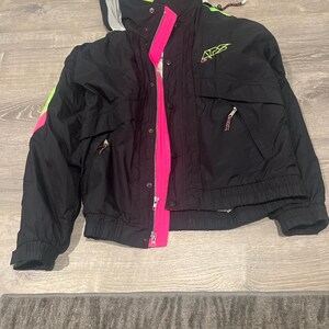May include: Black and neon green winter jacket with a pink zipper and accents. The jacket has a hood and two front pockets. The logo "APS" is visible on the right chest area. The jacket is made of a water-resistant material.