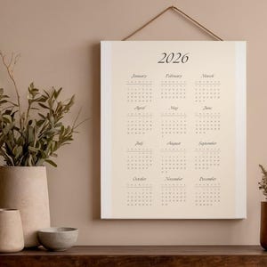 Op de afbeelding: Een crèmekleurige 2026-kalender met een minimalistisch ontwerp, opgehangen aan een bruin touwtje. De kalender toont de maanden en data in een eenvoudig, elegant lettertype. De achtergrond is neutraal beige.