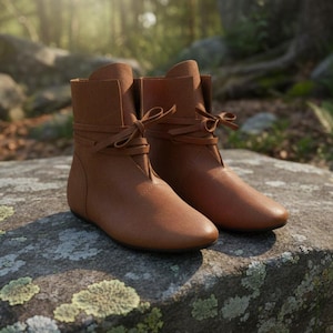 May include: Brown leather ankle boots with a lace-up detail. The flat-soled boots are displayed on a mossy rock. The boots have a soft, matte finish. The background is a blurred natural scene with trees and sunlight.