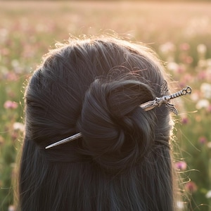 Puede incluir: Una horquilla plateada con forma de espada y alas detalladas en la empuñadura, insertada horizontalmente en un moño de pelo oscuro. El accesorio de metal presenta un diseño intrincado en el mango y una hoja afilada y puntiaguda.