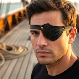 May include: Black leather eye patch with a compass rose design, secured with a black strap and gold-colored rivets. The eye patch is worn over one eye. The background shows a wooden deck and rigging.