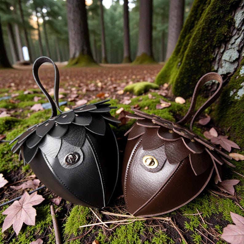 May include: Two acorn-shaped purses, one black and one brown, rest on mossy ground. Each purse features a petal-like design around the top and a small handle. The purses have a metal clasp closure. The background shows a forest with tall trees.
