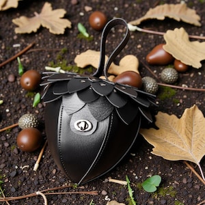 May include: A black leather handbag shaped like an acorn, with a handle and a decorative flap design. The bag features a silver clasp and is surrounded by acorns and fall leaves.