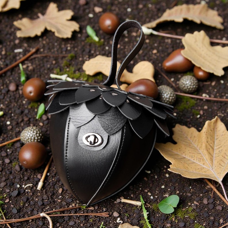 May include: A black leather handbag shaped like an acorn, with a handle and a decorative flap design. The bag features a silver clasp and is surrounded by acorns and fall leaves.