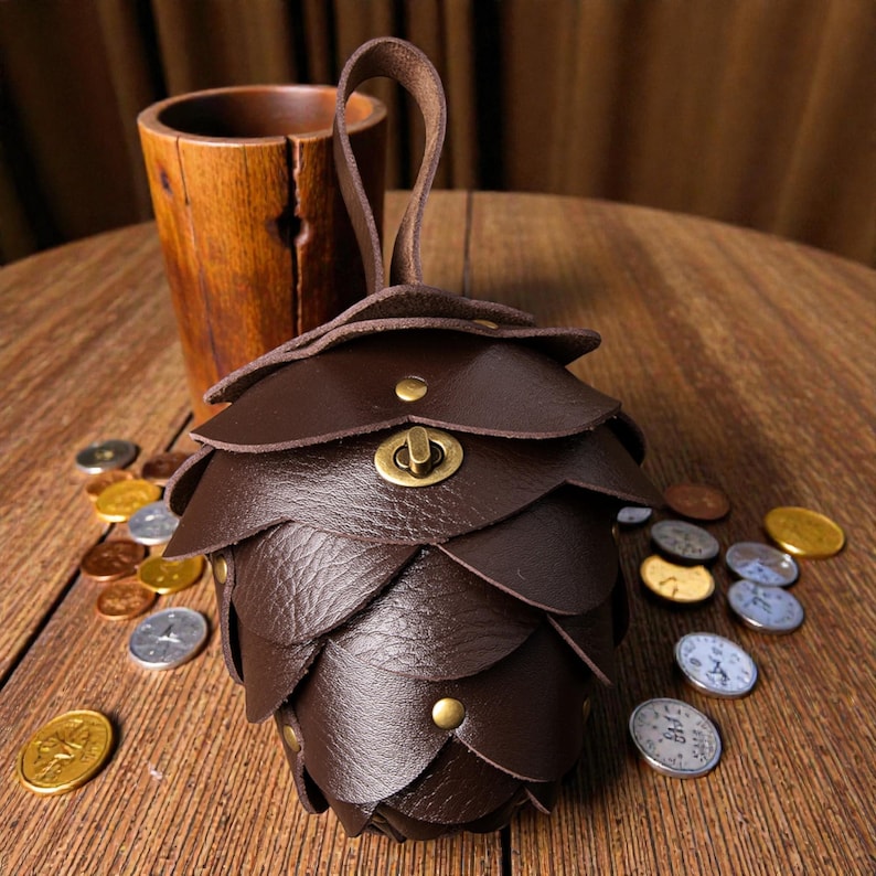 May include: A handcrafted brown leather coin purse shaped like a pinecone, with a small handle and brass hardware. The purse is surrounded by various coins on a wooden surface. A wooden cup is in the background.