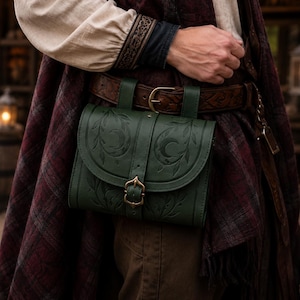 May include: A dark green leather belt pouch featuring an embossed crescent moon and leaf pattern on the front flap. The bag is secured with a brass-toned metal buckle and is attached to a wide, brown, tooled leather belt worn around the waist.