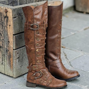 Puede incluir: Botas altas de cuero marrón con múltiples hebillas y correas. Las botas tienen una estética vintage, steampunk, con detalles metálicos decorativos y un acabado ligeramente desgastado. Las botas están junto a una caja de madera.