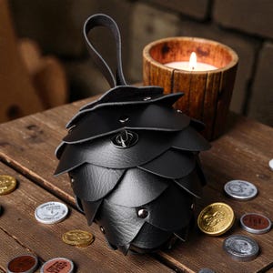 May include: Black leather coin purse shaped like a pinecone, with a loop handle and metal accents. The purse is surrounded by coins on a wooden surface, with a lit candle in a wooden holder in the background.