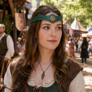 May include: A green leather headband featuring a central gold-toned circular emblem with a deer motif and black vine-like detailing. The headband is worn across the forehead, complemented by a dark cord necklace with a textured leaf-shaped pendant.