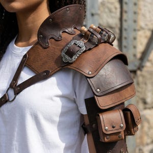 May include: A brown leather shoulder armor with a decorative silver buckle and small pouches. The armor has multiple layers and is designed to protect the shoulder and upper arm. Small glass vials are attached to the armor.