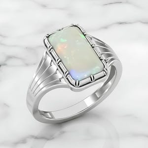 Op de afbeelding: Zilveren ring met een rechthoekige opaal edelsteen. De opaal heeft een melkachtige witte basis met iriserende kleuren. De ring heeft een gedetailleerde zetting en een gepolijste band. De ring staat op een witte marmeren achtergrond.