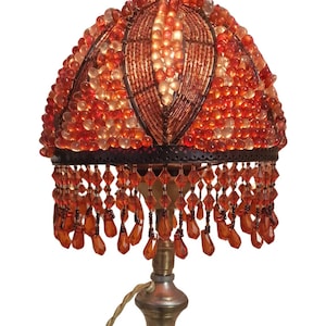 Puede incluir: Lámpara de mesa vintage con una pantalla abovedada cubierta de cuentas naranjas y ámbar. La pantalla está adornada con cuentas en forma de lágrima. La lámpara tiene una base de latón con un diseño ornamentado y una base circular negra. Un cable dorado sale de la lámpara.