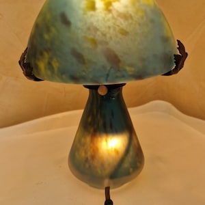 May include: A mushroom-shaped table lamp with a teal and yellow glass shade. The lamp has a dark brown base and a matching cord. The lamp is lit, casting a warm glow.