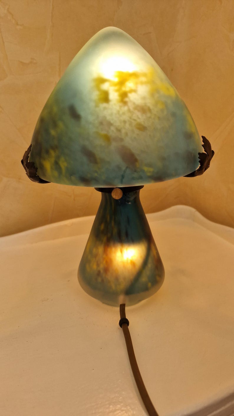 May include: A mushroom-shaped table lamp with a teal and yellow glass shade. The lamp has a dark brown base and a matching cord. The lamp is lit, casting a warm glow.