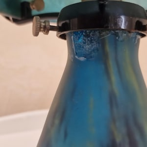 May include: Close-up of a blue glass lamp base with a flared shape. The base has a gradient of blue and green hues. The lamp's hardware includes brass-colored screws and a black metal fixture.