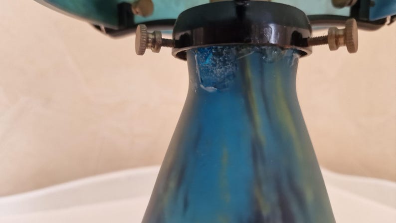 May include: Close-up of a blue glass lamp base with a flared shape. The base has a gradient of blue and green hues. The lamp's hardware includes brass-colored screws and a black metal fixture.
