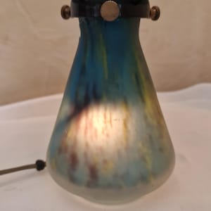 May include: A vintage table lamp with a blue, yellow, and brown glass base. The lamp has a black metal frame and a light blue lampshade. The lamp is lit, and the base has a unique, colorful design.