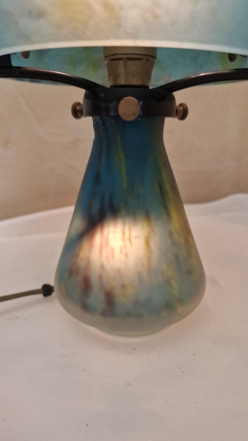 May include: A vintage table lamp with a blue, yellow, and brown glass base. The lamp has a black metal frame and a light blue lampshade. The lamp is lit, and the base has a unique, colorful design.