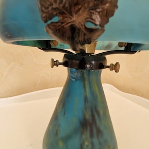 May include: A vintage-style table lamp with a teal glass shade and base. The shade features a dark brown leaf design. The lamp has brass accents and a black base. The lamp is on a white surface.