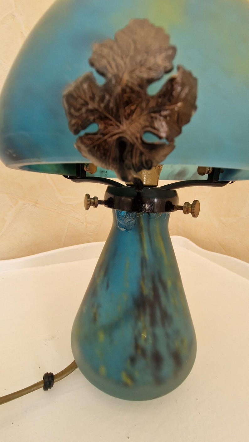 May include: A vintage-style table lamp with a teal glass shade and base. The shade features a dark brown leaf design. The lamp has brass accents and a black base. The lamp is on a white surface.