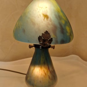 May include: A mushroom-shaped table lamp with a glass shade. The shade is a blend of blue, yellow, and white, with a leaf-shaped accent. The base is a darker teal color. The lamp is lit, creating a warm glow.
