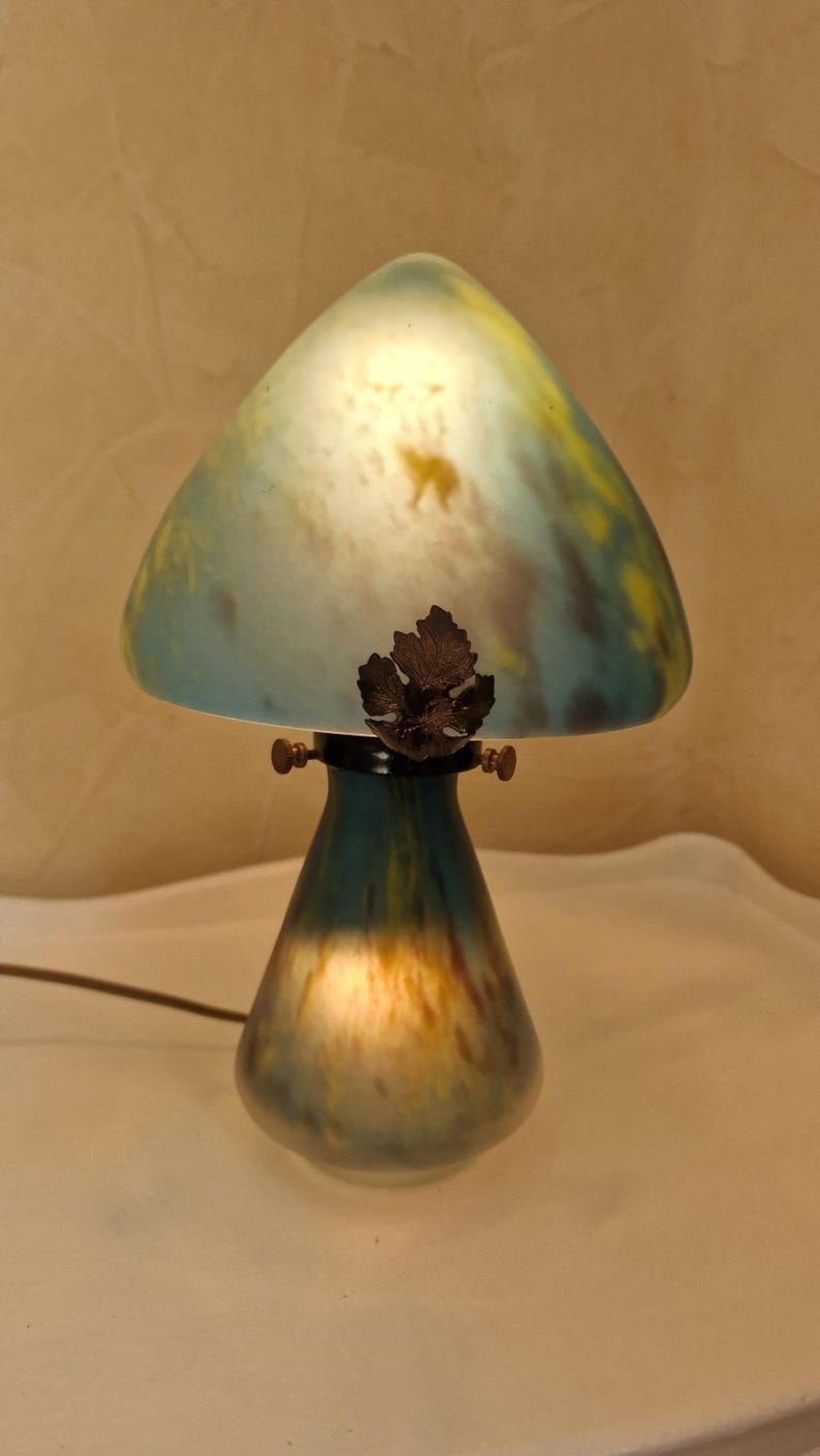 May include: A mushroom-shaped table lamp with a glass shade. The shade is a blend of blue, yellow, and white, with a leaf-shaped accent. The base is a darker teal color. The lamp is lit, creating a warm glow.