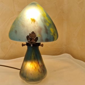May include: A mushroom-shaped table lamp with a glass shade. The shade is a mix of blue, yellow, and green, with a leaf-shaped accent. The base is a dark teal color. The lamp is on a white surface.